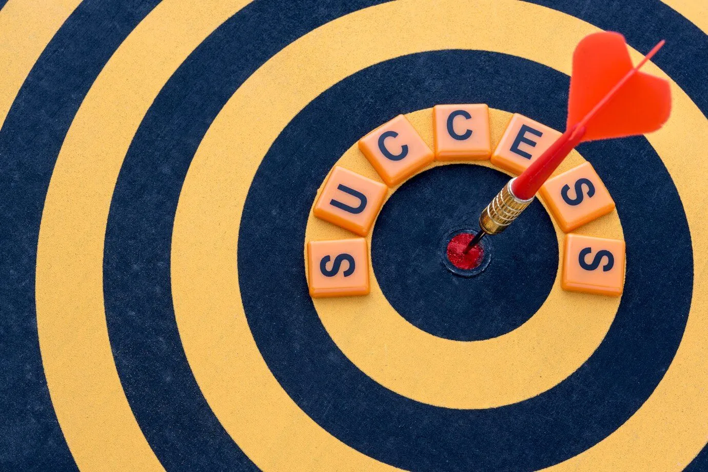 A dart in the bullseye of a dartboard, ringed by tiles spelling the word success