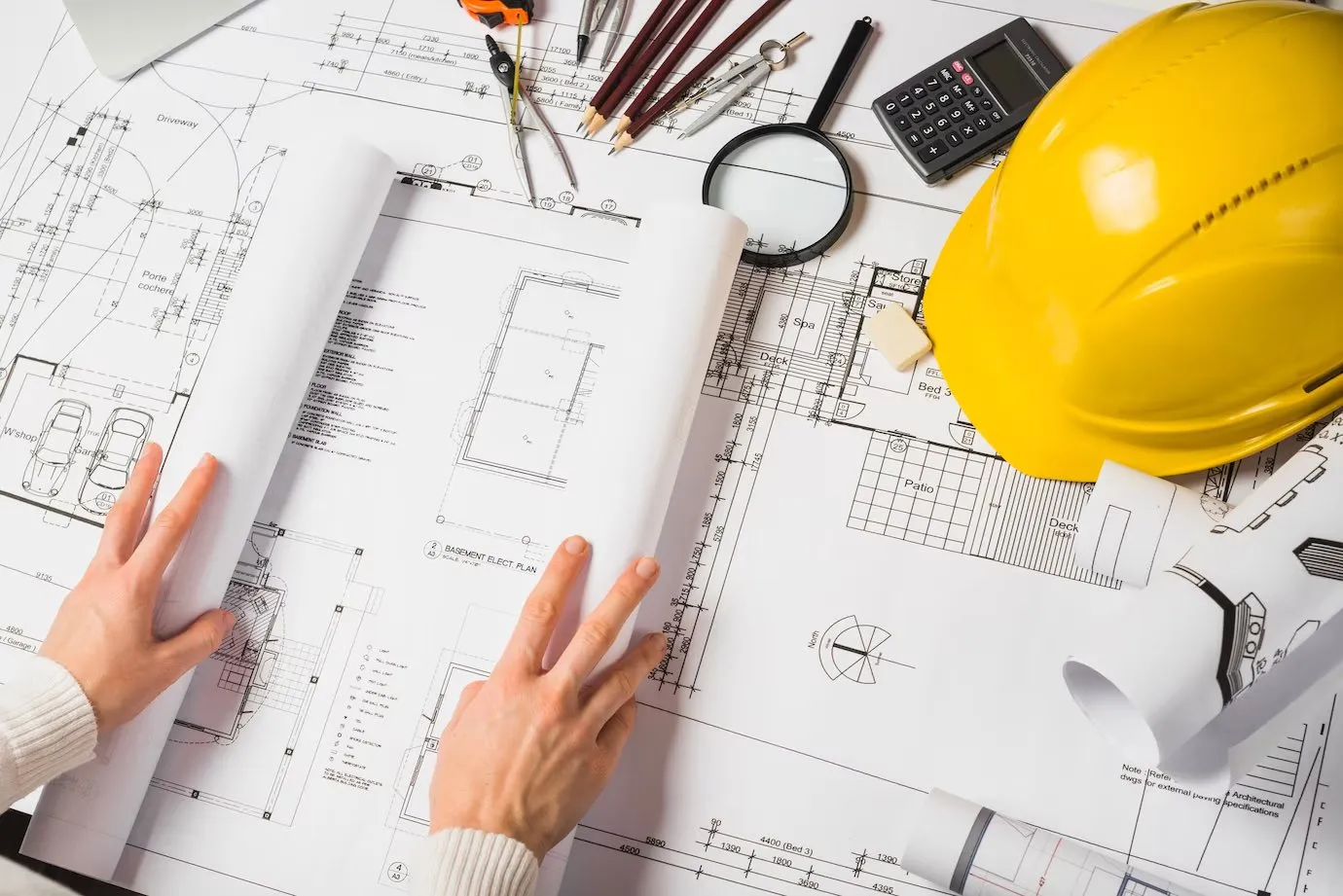 Hands spread over architectural plans beside a yellow hard hat and drawing tools