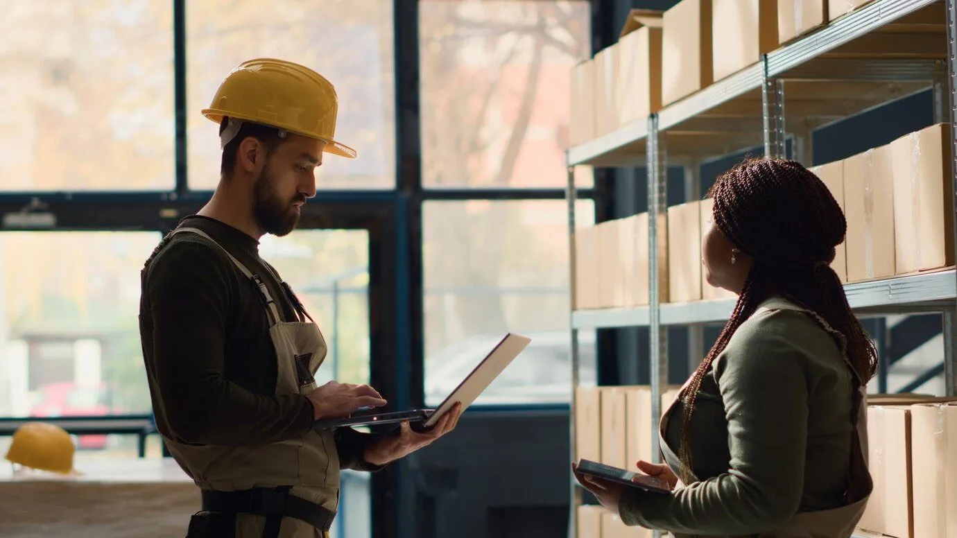 A worker in a hard hat holding a laptop while talking to a colleague among storage shelving