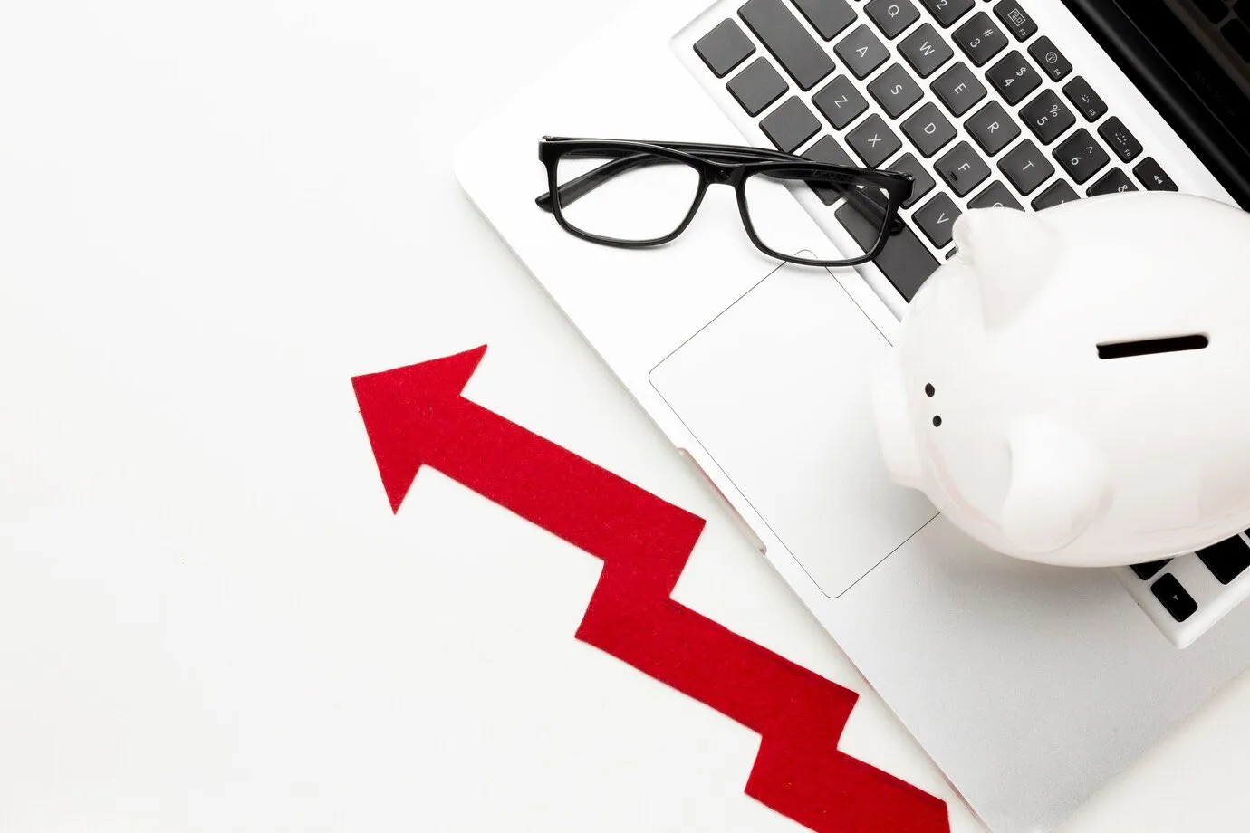 A red upward arrow beside a piggy bank and glasses resting on a laptop