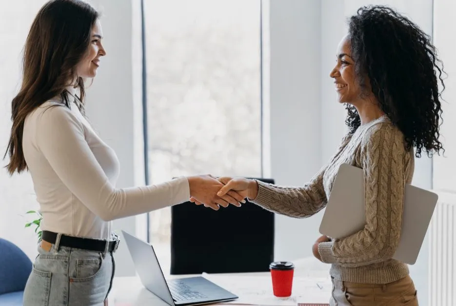 Two people shaking hands across a desk in a bright office