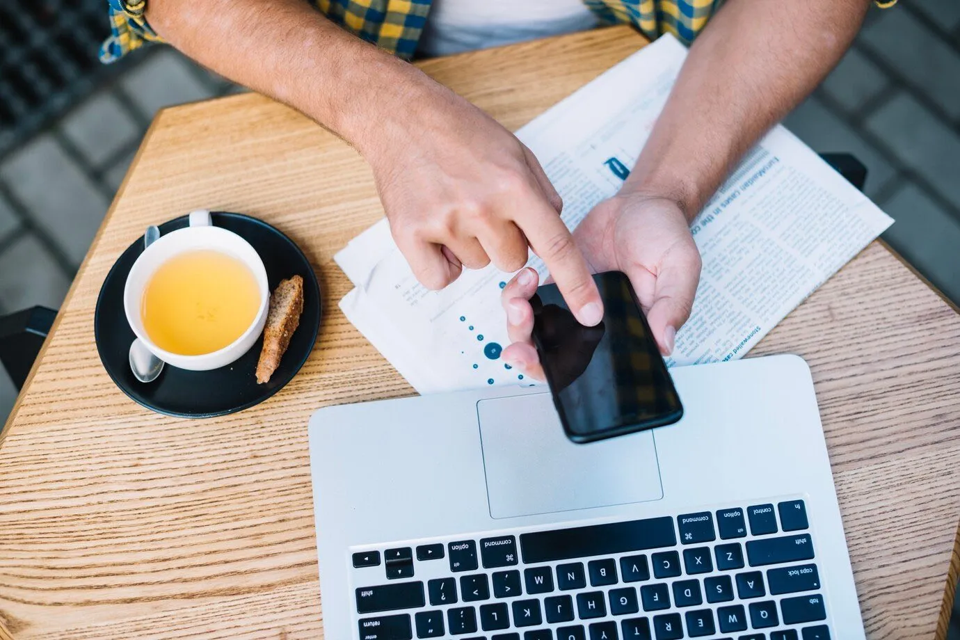A person using a smartphone over an open laptop at a cafe table