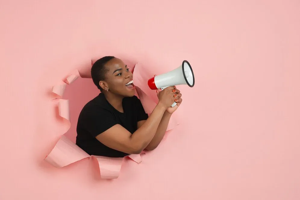 A woman calling out through a megaphone from a torn hole in pink paper